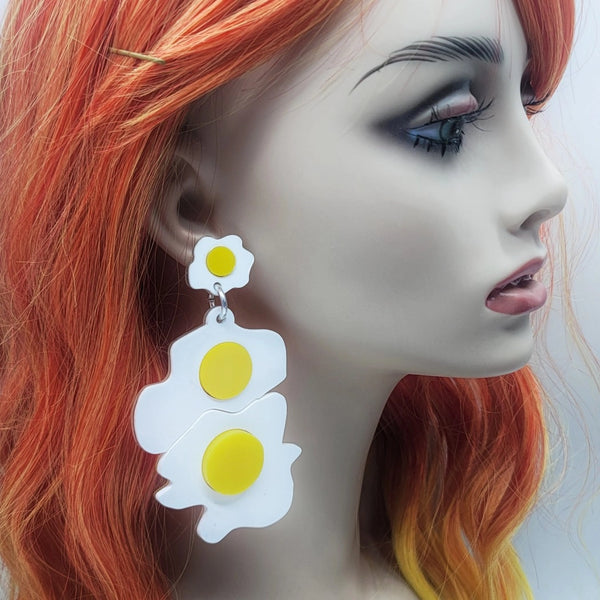 A pair of laser cut acrylic drop earrings in the shape of a rare ribeye steak with marbled red acrylic and two sunny side up eggs. Eggs are shown on the ear of a mannequin 