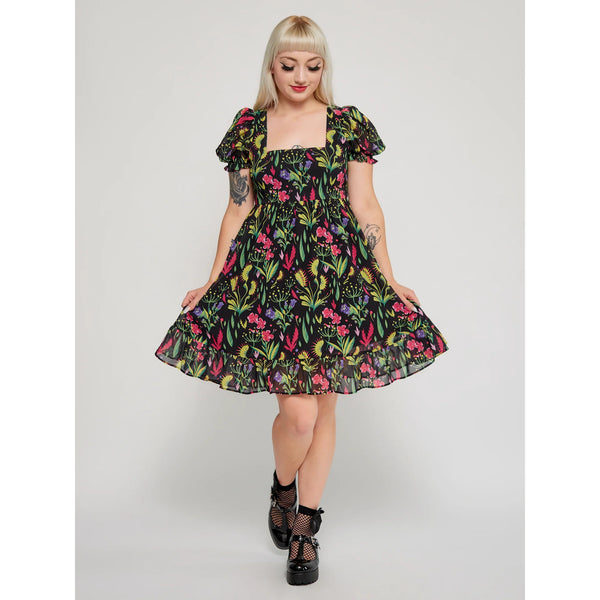 Model wearing a mini dress with a botanical pattern including Venus Flytraps on a black background. It has a square neckline, slightly puffed short sleeves, and a full skirt with a ruffled hem. Shown from the front with skirt held wide