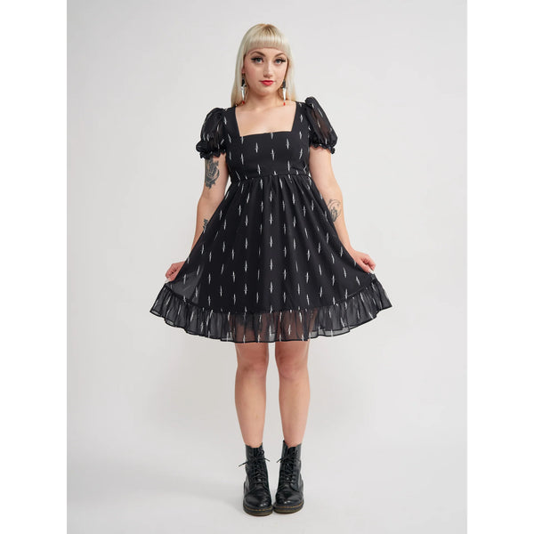Model wearing a black mini dress with an all-over tiny pattern of white traditional tattoo style daggers. It has a square neckline, slightly puffed short sleeves, and a full skirt with a ruffled hem. Shown from the front in a full length shot with skirt held wide