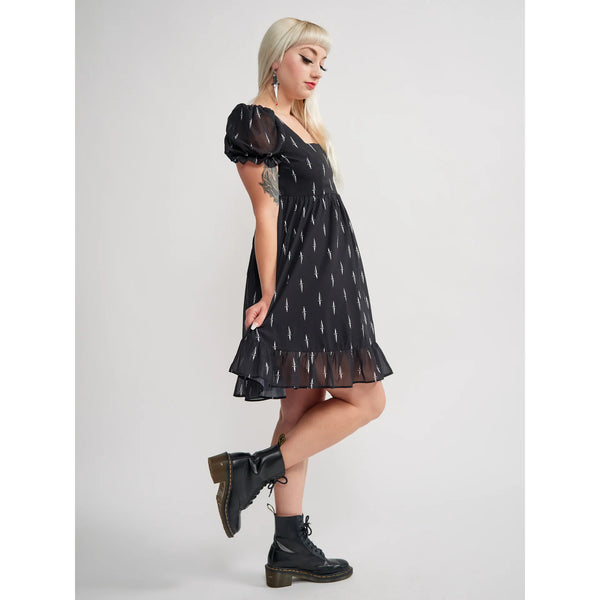 Model wearing a black mini dress with an all-over tiny pattern of white traditional tattoo style daggers. It has a square neckline, slightly puffed short sleeves, and a full skirt with a ruffled hem. Shown from a three quarter angle