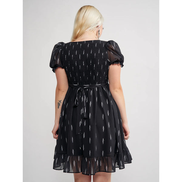 Model wearing a black mini dress with an all-over tiny pattern of white traditional tattoo style daggers. It has a square neckline, slightly puffed short sleeves, and a full skirt with a ruffled hem. Shown from behind