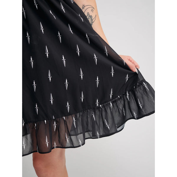 Model wearing a black mini dress with an all-over tiny pattern of white traditional tattoo style daggers. It has a square neckline, slightly puffed short sleeves, and a full skirt with a ruffled hem. Shown in close up to display pattern detail
