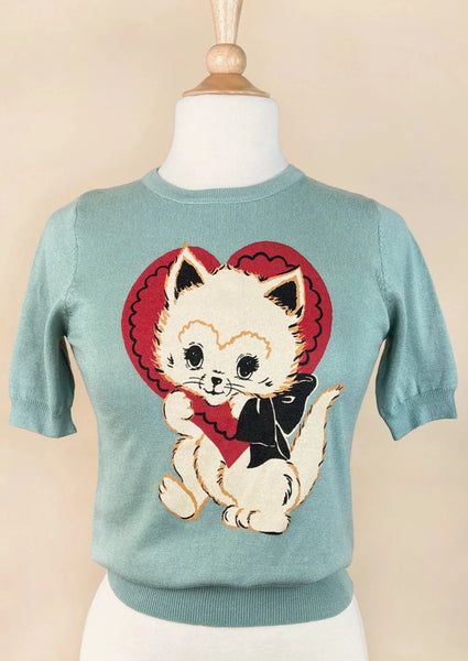 a powder blue short sleeved sweater with a printed illustration of a white kitten with a red valentine heart around its head and a black bow around its neck. Shown on a dress form