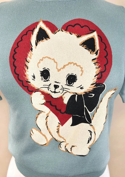a powder blue short sleeved sweater with a printed illustration of a white kitten with a red valentine heart around its head and a black bow around its neck. Shown on a dress form in close up