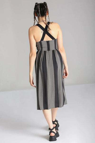 A model wearing a sleeveless dress in a grey and black checker plaid pattern with alternating wide black vertical straps. It has wide adjustable self straps with O-ring detail connecting them to the dress. The dress has princess seaming, a shallow scoop neckline, and a full gathered skirt that ends below the knee. Shown from the back