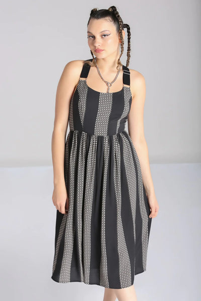 A model wearing a sleeveless dress in a grey and black checker plaid pattern with alternating wide black vertical straps. It has wide adjustable self straps with O-ring detail connecting them to the dress. The dress has princess seaming, a shallow scoop neckline, and a full gathered skirt that ends below the knee. Shown from the front