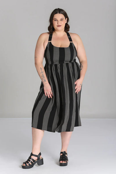A plus size model wearing a sleeveless dress in a grey and black checker plaid pattern with alternating wide black vertical straps. It has wide adjustable self straps with O-ring detail connecting them to the dress. The dress has princess seaming, a shallow scoop neckline, and a full gathered skirt that ends below the knee. Shown from the front 