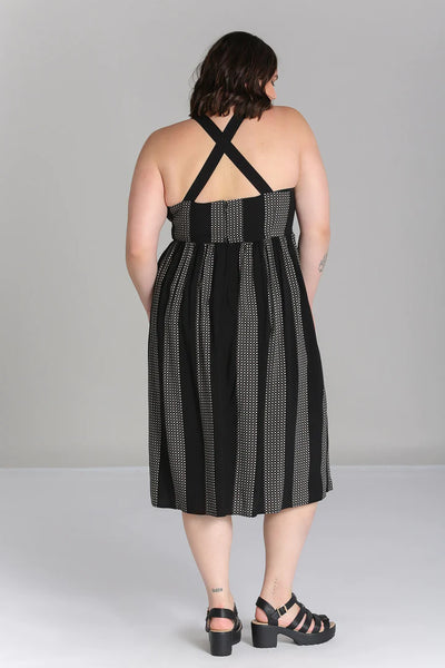 A plus size model wearing a sleeveless dress in a grey and black checker plaid pattern with alternating wide black vertical straps. It has wide adjustable self straps with O-ring detail connecting them to the dress. The dress has princess seaming, a shallow scoop neckline, and a full gathered skirt that ends below the knee. Shown from the back
