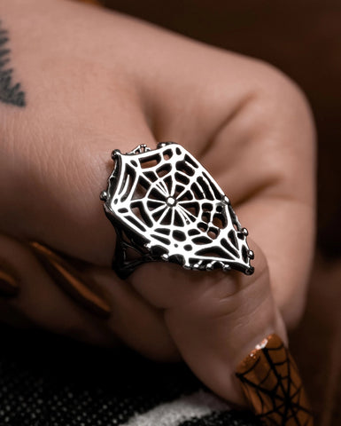 Model wearing stainless steel ring on their thumb in the shape of a shield-like spider web with openwork design and scalloped edges. Shown in close up
