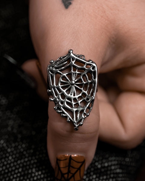 Model wearing stainless steel ring on their thumb in the shape of a shield-like spider web with openwork design and scalloped edges. Shown in close up from a straight ahead angle