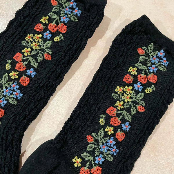 Black cotton socks with both a textured cable knit-in pattern and knit-in pattern of tiny strawberries and yellow & blue flowers. Shown in close up