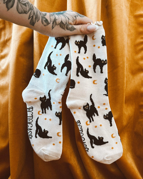 Pair of off-white crew socks with ruffle cuff and all-over pattern of black cats with gold crescent moons and stars. Ectogasm logo on bottom of sock. Held by model in front of a golden colored velvet curtain