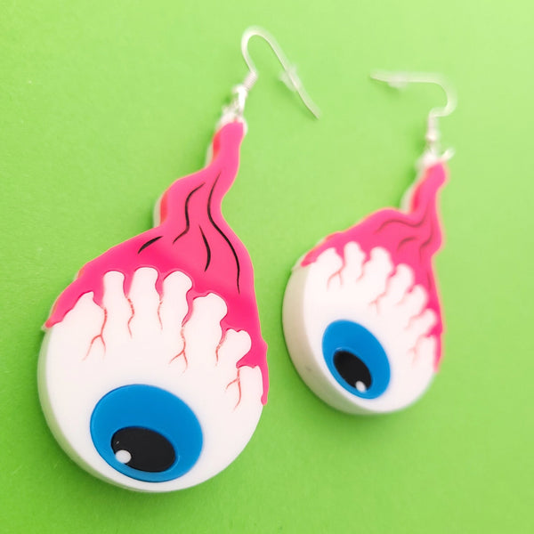 A pair of laser-cut acrylic dangle earrings of a pair of cartoony bloodshot eyeballs with blue irises hanging from bright pink optic nerves. Shown flat in close up
