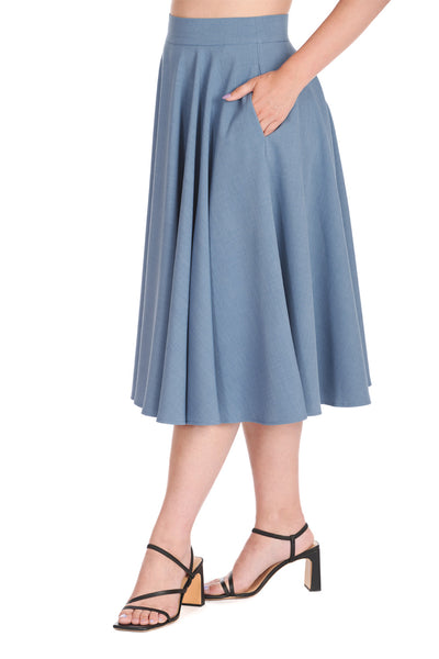 Model wearing a high-waisted swing skirt in a medium shade of cornflower blue that ends just below the knee. Shown from a three quarter angle with hands in side seam pockets