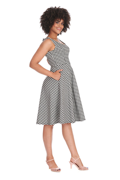 A model wearing a black and white gingham pattern sleeveless fit and flare dress. It has a slightly scooped neckline, princess seaming, side seam pockets, and a skirt that ends just below the knee. Shown from three quarter angle to display pockets