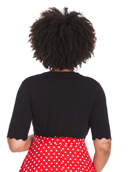 Model wearing a black bolero length open cardigan with elbow-length sleeves and a rounded bottom hem with wavy scalloped ends. Shown from the back