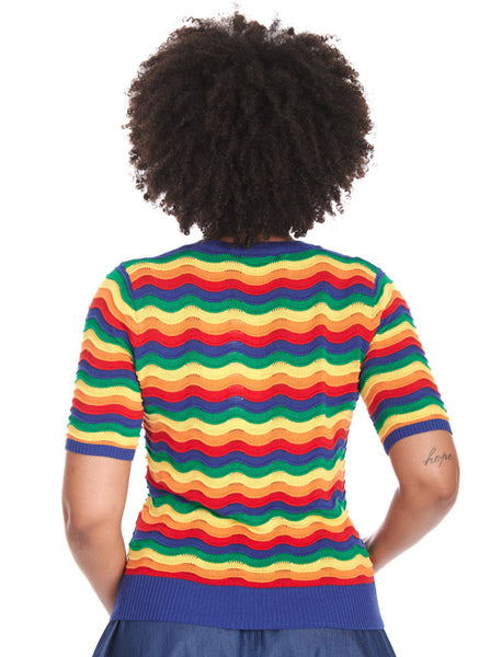 A model wearing a short sleeved sweater with a round neckline and elbow length sleeves in an all over knit in wavy rainbow pattern with small pointelle details at the top of each wave. The sweater has bright blue ribbed collar, cuffs, and bottom band. Shown from the back untucked