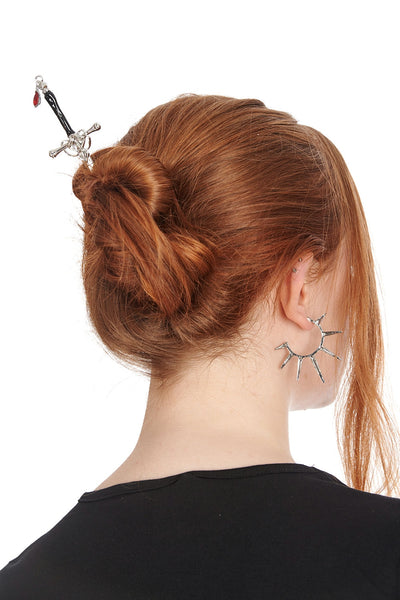 Silver metal hair stick in the shape of a sword with orange silver and black handle and red glass bead in the shape of a drop of blood at the end of the handle. Shown in the updo of a model