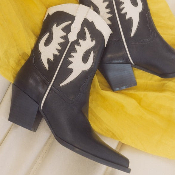 A pair of black pointed toe western style boots with pointed toes and 2” block heels. Black shiny faux leather with creamy white inset details. Shown from above on their sides