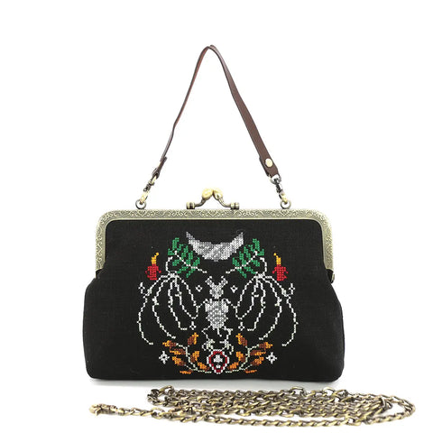 Kiss lock style purse with antiqued bronze metal hardware and rectangular black fabric body. Has embroidered needlepoint style decoration of a bat’s skeleton surrounded by burning candles, leaves, and a crescent moon and stars. Purse has a brown faux leather hand strap and a link style chain shoulder strap 