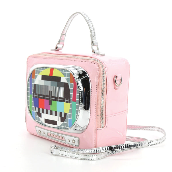 A rectangular novelty purse in the shape of a vintage television set in shiny pastel pink vinyl with silver details. Has a “screen” with a colorful test pattern and faux knob controls. Has a silver handle and detachable matching cross body strap. Shown from a three quarter angle to display the three sided zipper