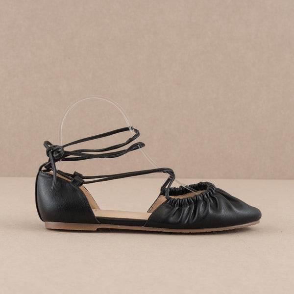 A pair of black faux leather ballet flats with matching faux leather laces running along the edge of the vamp, crisscrossing to the sides of each ankle, and ending at the back of the heel. Shown unworn from the side 