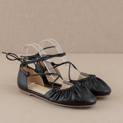 A pair of black faux leather ballet flats with matching faux leather laces running along the edge of the vamp, crisscrossing to the sides of each ankle, and ending at the back of the heel. Shown unworn from a three quarter angle