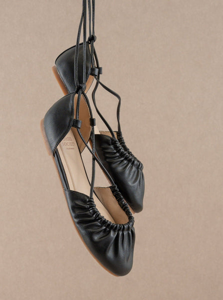 A pair of black faux leather ballet flats with matching faux leather laces running along the edge of the vamp, crisscrossing to the sides of each ankle, and ending at the back of the heel. Shown hanging