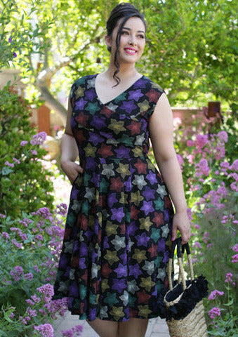 A model wearing a fit and flare style dress in a cotton spandex knit with an all over pattern of fuchsia, purple, red, orange, creamy off-white, and teal blooming tropical flowers on a black background. Dress is faux-wrap style with a surplice bodice and Queen Anne neckline. Full skirt ends below the knee and has side seam pockets. Seen from front
