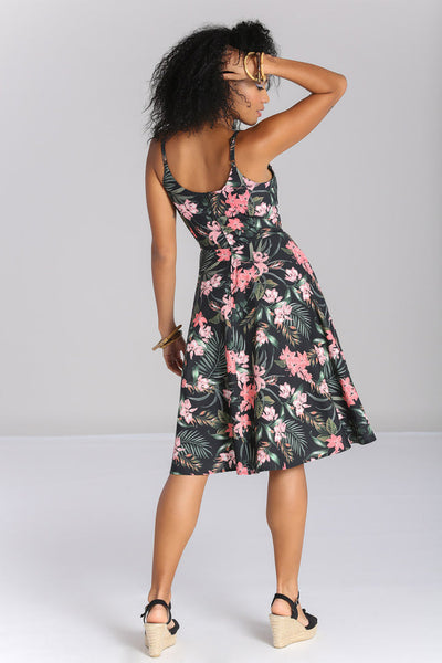 Model wearing a sleeveless 1950’s style sundress in a stretch cotton fabric. All-over print on a black background of pink tropical flowers and green foliage. Dress has a sweetheart neckline, princess seaming, adjustable thin straps, and a full below the knee skirt with pockets. Seen from the back 