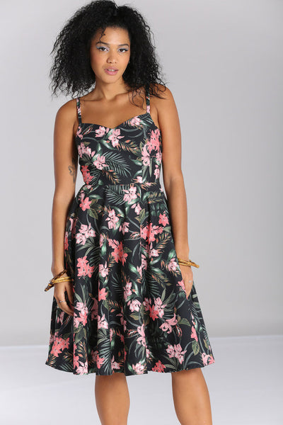 Model wearing a sleeveless 1950’s style sundress in a stretch cotton fabric. All-over print on a black background of pink tropical flowers and green foliage. Dress has a sweetheart neckline, princess seaming, adjustable thin straps, and a full below the knee skirt with pockets. Seen from the front