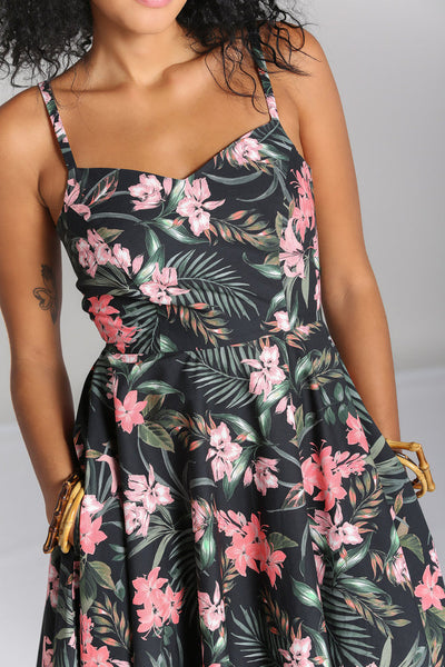 Model wearing a sleeveless 1950’s style sundress in a stretch cotton fabric. All-over print on a black background of pink tropical flowers and green foliage. Dress has a sweetheart neckline, princess seaming, adjustable thin straps, and a full below the knee skirt with pockets. Seen from the front in close up