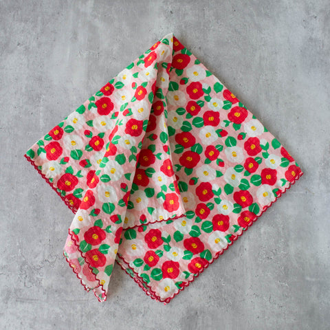 Cotton square scarf with allover print of Camellia flowers in red and white with yellow pistils and green leaves on a light pink background. Scarf has scalloped red border