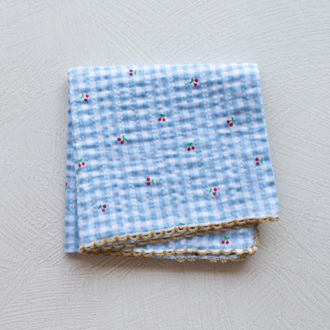 Cotton square scarf with allover blue and white gingham pattern & tiny red and green cherry print. Edges of scarf have light brown embroidered scalloped edge. Shown folded