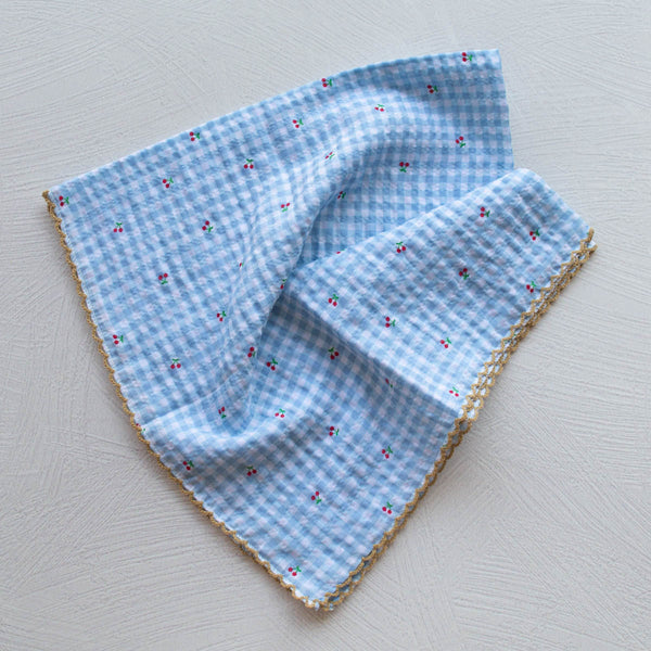 Cotton square scarf with allover blue and white gingham pattern & tiny red and green cherry print. Edges of scarf have light brown embroidered scalloped edge. Shown unfolded