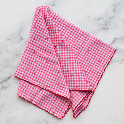 Square cotton scarf in a medium pink and white gingham pattern and tiny red and green cherry print. Scarf has a deep red scalloped embroidered edge. Shown unfolded