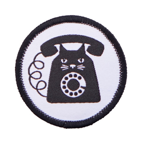 A woven round patch with embroidered black edge featuring an image of a black and white cat with rotary phone features on a white background 