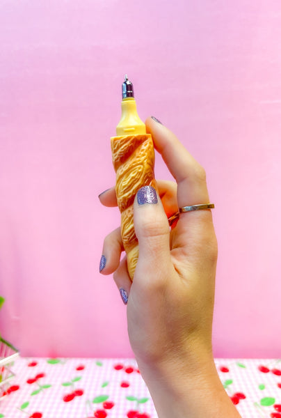 A novelty ballpoint pen with an exterior shape of a loaf of baguette bread. Shown open held by a model on a pink background 