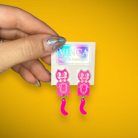 Hot pink drop earrings in the shape of small Kit Kat Klocks with moving tails. Shown on their cardboard backing held by hand in front of a yellow background 