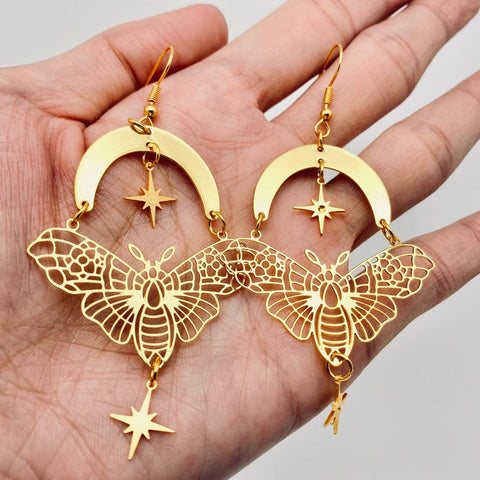 Shiny gold metal dangle earrings made of a sideways crescent moon, a starburst, and a large moth with cutout details connected by jump rings