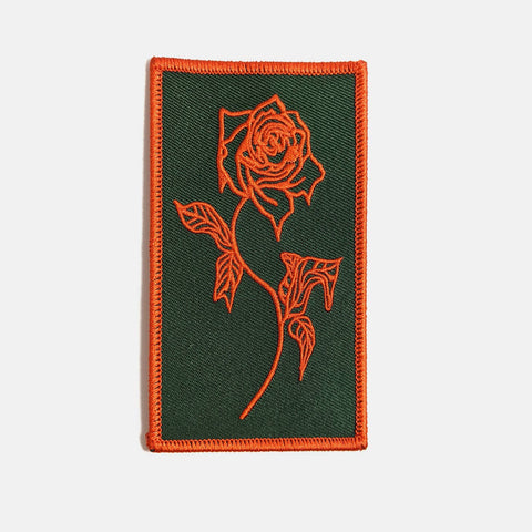 Rectangular embroidered patch of a large orange rose with stem and leaves that has glitched/melting details on a forest green background and matching orange border. Shown flat