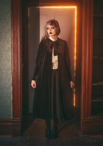 Model wearing black velvet duster with bow tied detail at the collar, slightly gathered shoulders, and Juliet sleeves. Shown from the front 
