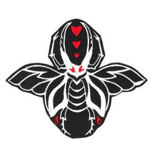 An embroidered patch of a large white beetle with wings on a black background. It has red eyes and there are three small red hearts in between its pincers. Shown flat