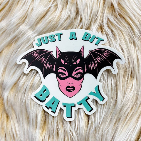 Die cut vinyl sticker of a pink retro looking woman wearing a black bat mask with wings and a head. Caption in mint blue “JUST A BIT BATTY”