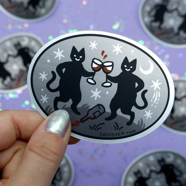 Oval vinyl sticker of two black cats holding glasses of red wine with a spilled bottle at their feet. On a metallic grey background with white moons and stars