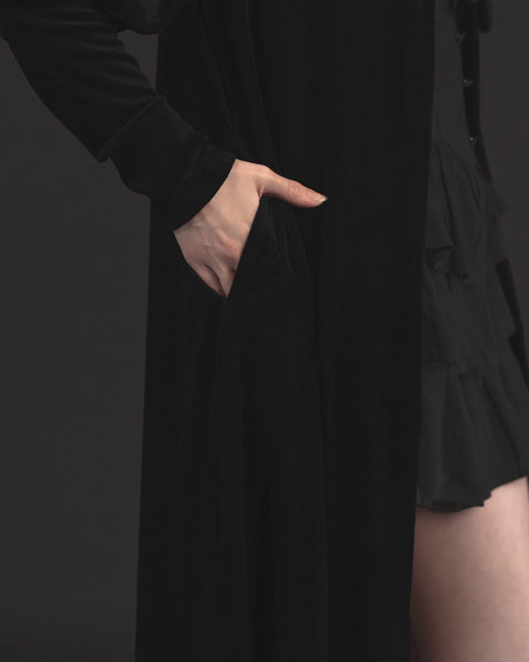 Model wearing black velvet duster with bow tied detail at the collar, slightly gathered shoulders, and Juliet sleeves. Showing detail of hand in side seam pocket