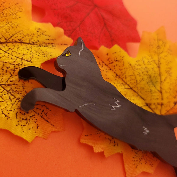A laser-cut acrylic brooch in a rippled black pattern of a black cat with painted silver detailing. Cat is pictured with limbs out and body extended. Yellow jewel detail for eye. Shown flat on an orange autumn leaf background up close for detail