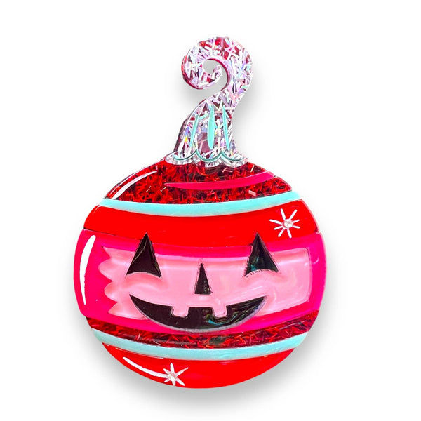 Spooktacular Sparkle Brooch by Johanna Parker x Lipstick & Chrome, tree ornament shaped brooch with black jack-o’-lantern face and green/red/pink stripes with red and iridescent glitter. Shown on white background 