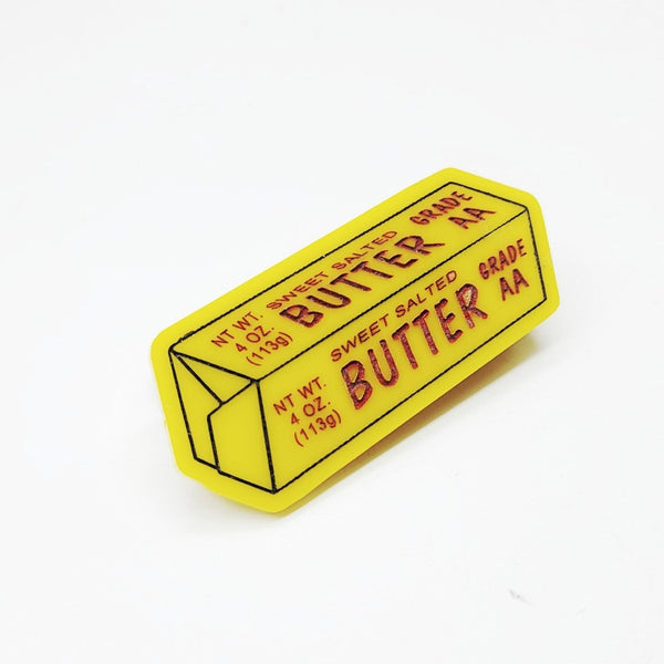 Laser-cut acrylic hair clip in the shape of a yellow stick of butter with black and red painted label details. Shown from the front 