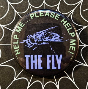 1.25” round button with purple image of fly on a black background with “HELP ME… PLEASE HELP ME!..” in green above the goth and “The Fly” below